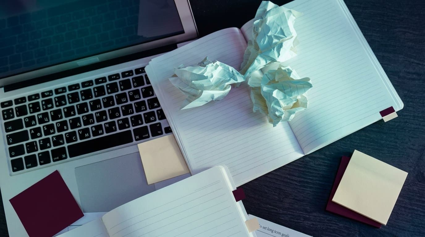 Overhead view of a laptop surrounded by crumpled paper, scattered notebooks, and sticky notes on a dark desk — representing the overwhelm of managing too many disconnected digital tools without a cohesive system.